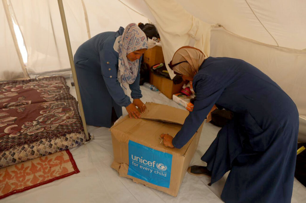 Enas and a neighbor open a box of hygiene supplies from UNICEF.
