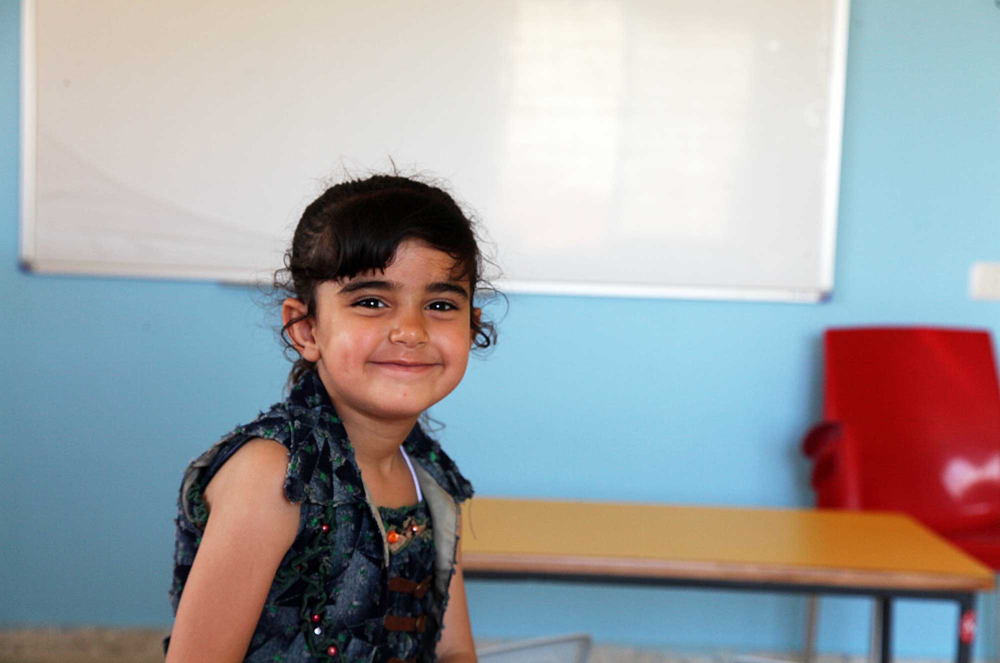 Raneen sits in her new classroom, smiling in her new dress. In the inset photo, Raneen stands in front of the ruined preschool during Anera’s psychosocial sessions directly following the war.