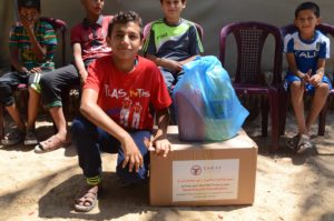 A young boy smiles while kneeling next to his food package. The gift will help his family eat healthy meals this Ramadan.