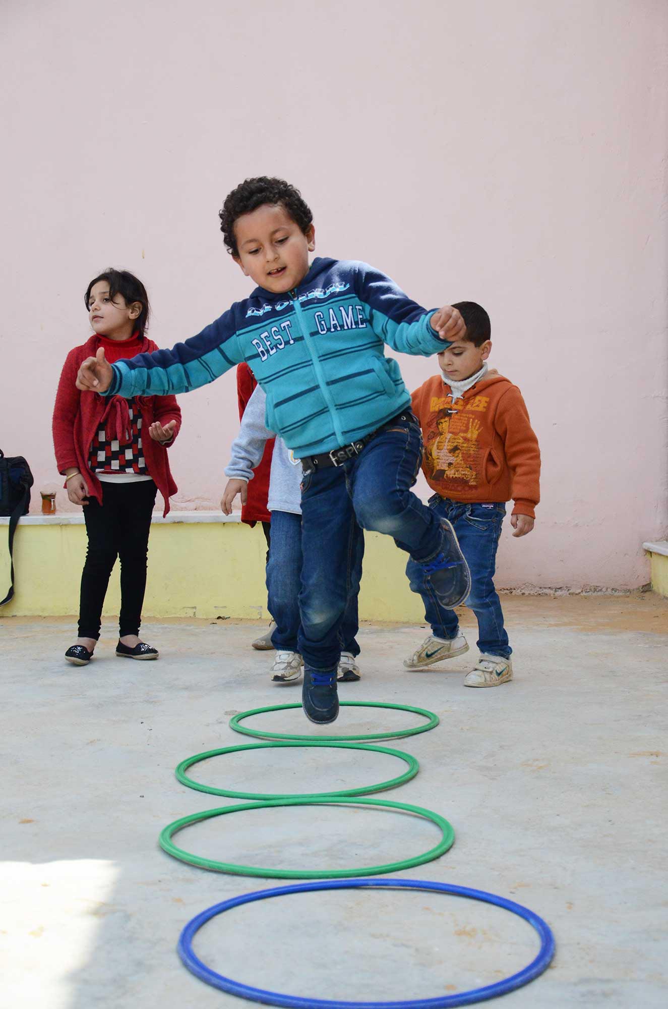 Nabil and his classmates take turns jumping into hoops during an outdoor play time.