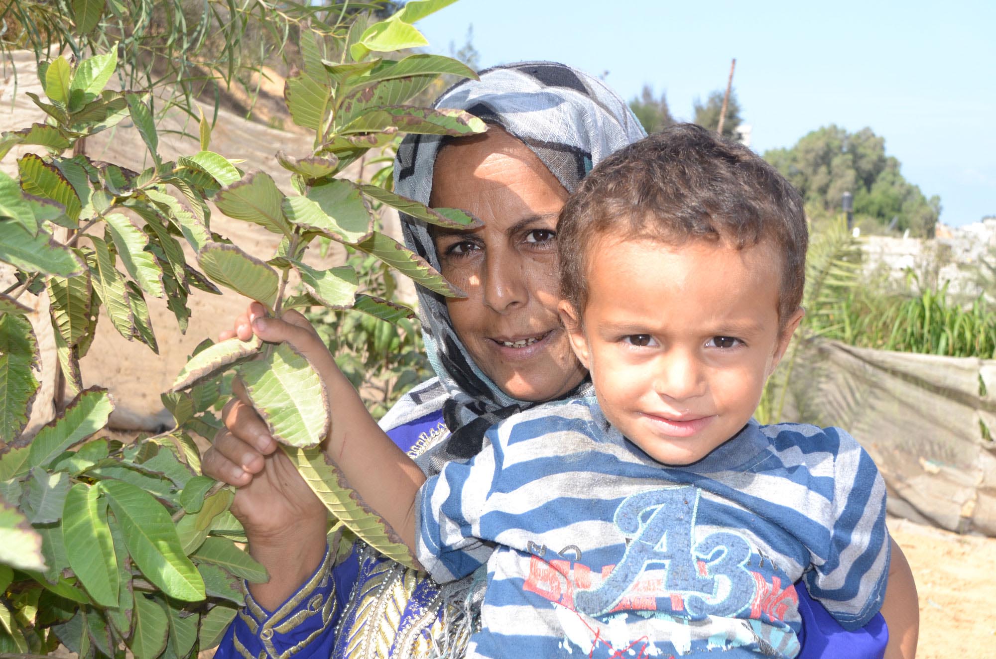 Amena El Masri and her grandchild in the shade of her Guava tree next to the new home garden Anera installed in her yard.