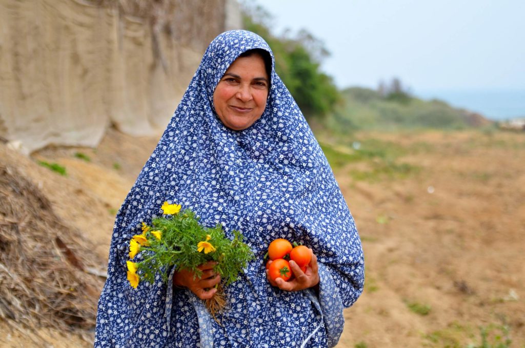 Amena standing on her land while showing off produce and flowers grown in her greenhouse.
