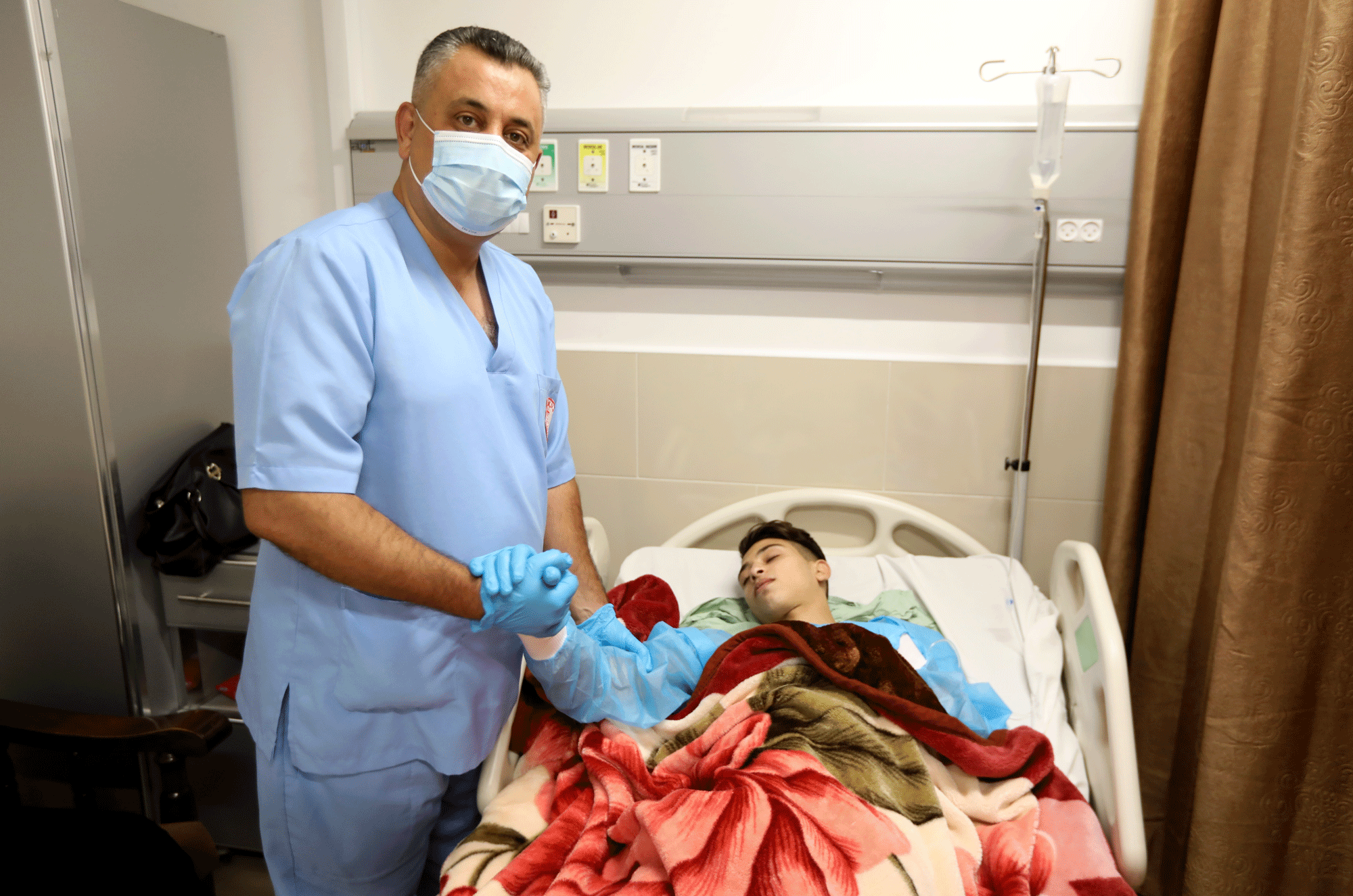 A gloved hospital employee holds a patient's hand.