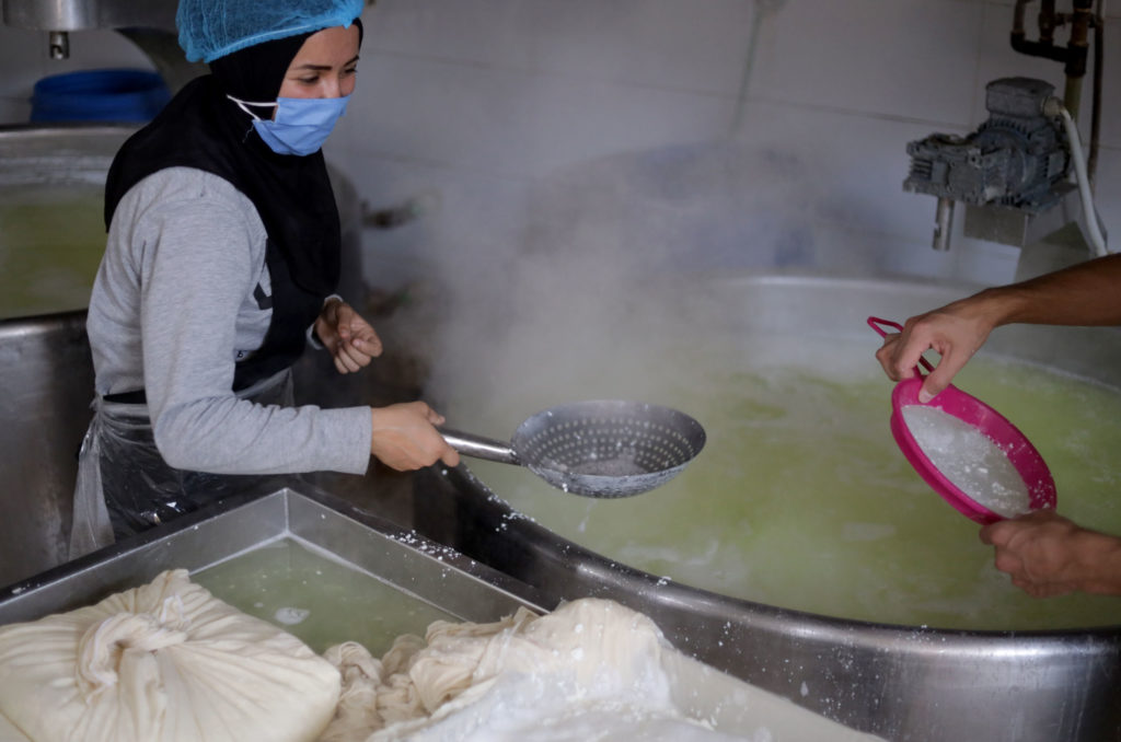 A trainee holds a strainer over a large vat of cheese water.