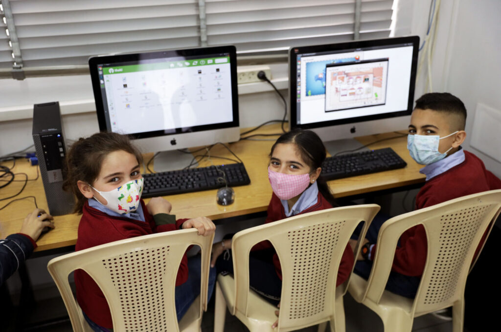 Three children sit at a table, sharing two Mac desktop computers. They are turned towards the camera and smiling.