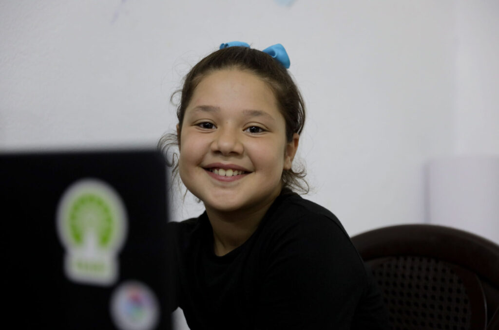 A girl faces the camera and smiles, her laptop obscures part of the frame. 