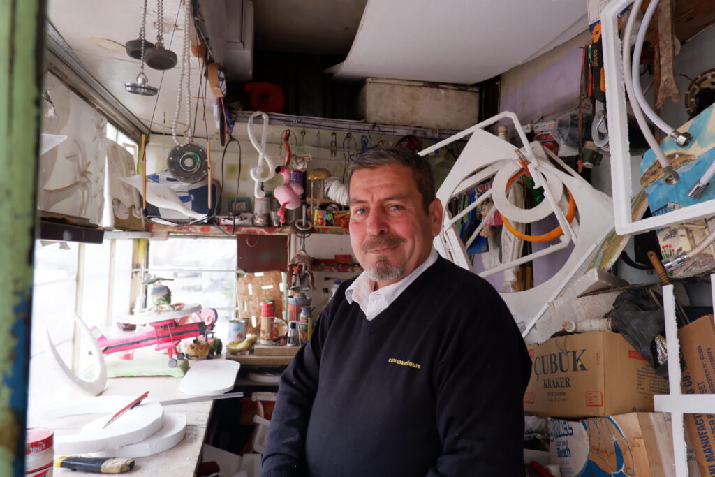 A man stands inside his artist studio surrounded from floor to ceiling by fragments of styrofoam sculpture.