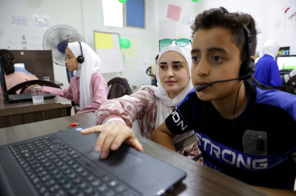 A boy wearing a headset faces his laptop and a woman sitting next to him leans over to use the trackpad.