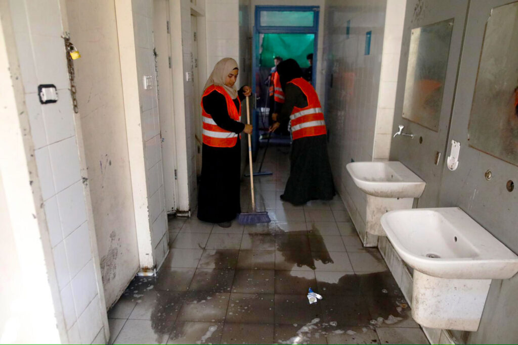 Two women mop a dirty bathroom floor in a shelter in Gaza.