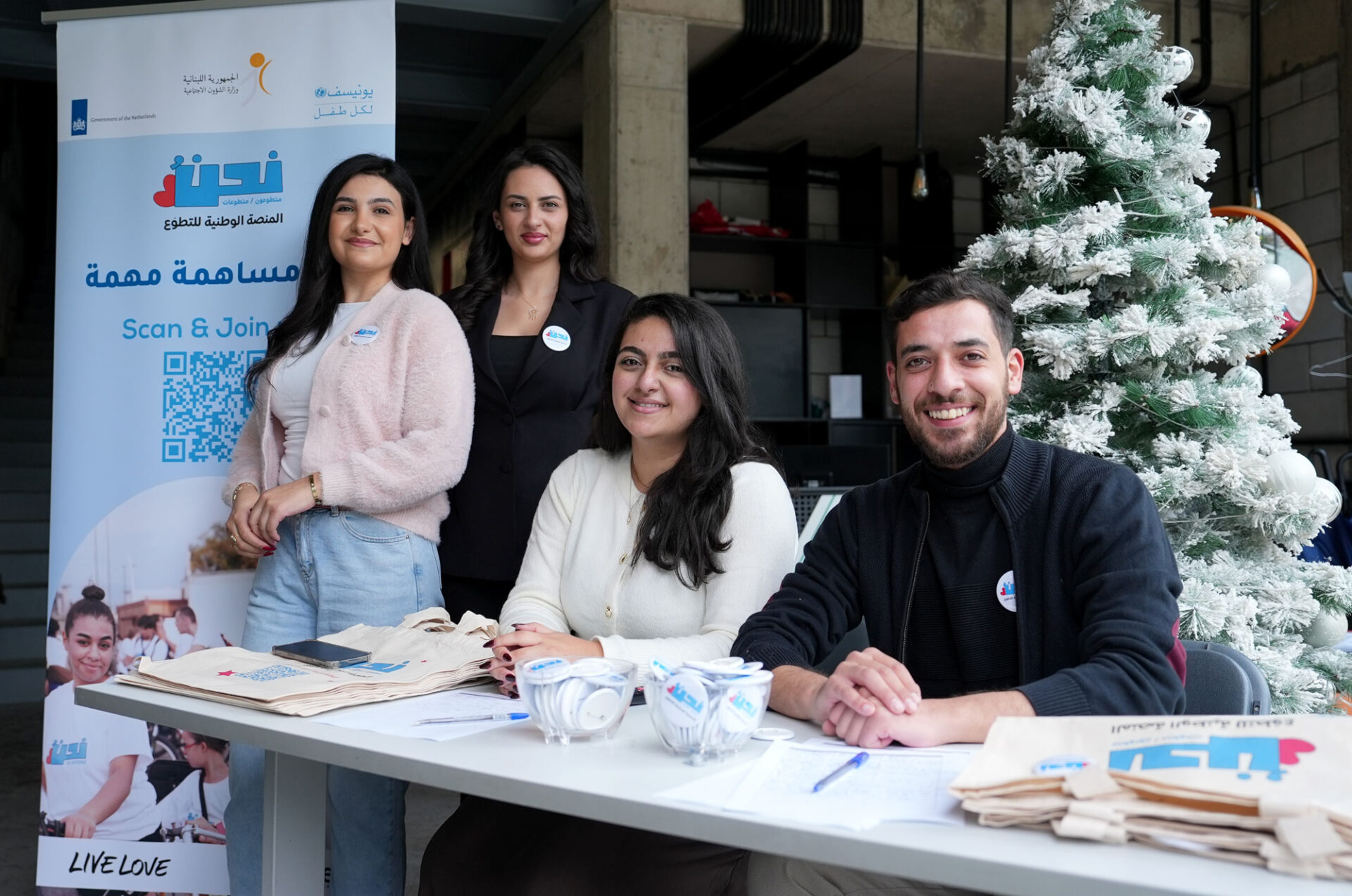 Young people staff a table with tote bags pins and a banner promoting the Nahno Volunteer program.