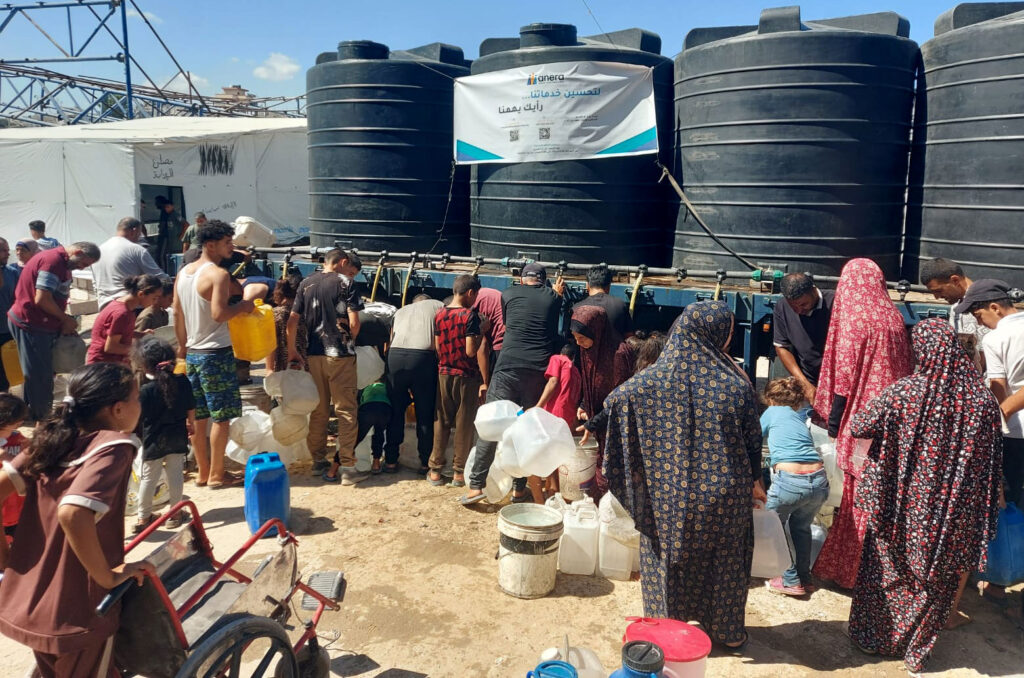 A crowd gathers for water with their containers at a water truck. A girl pushes a wheelchair that will carry the water jugs.
