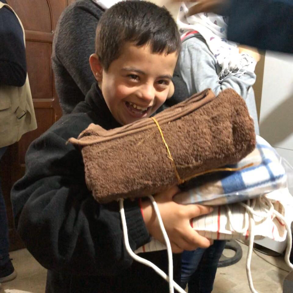 A young Syrian boy holds towles and blankets, made by Anera sewing course graduates and distributed by Anera in refugee camps in Lebanon.